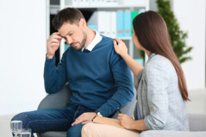 Depressed man looking at floor with hand on his forehead receiving counselling from female counselor