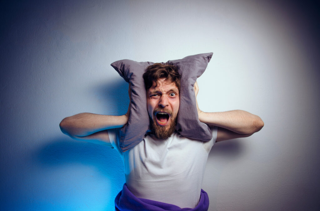 Man suffering from lack of sleep squishing a pillow against his ears and shouting